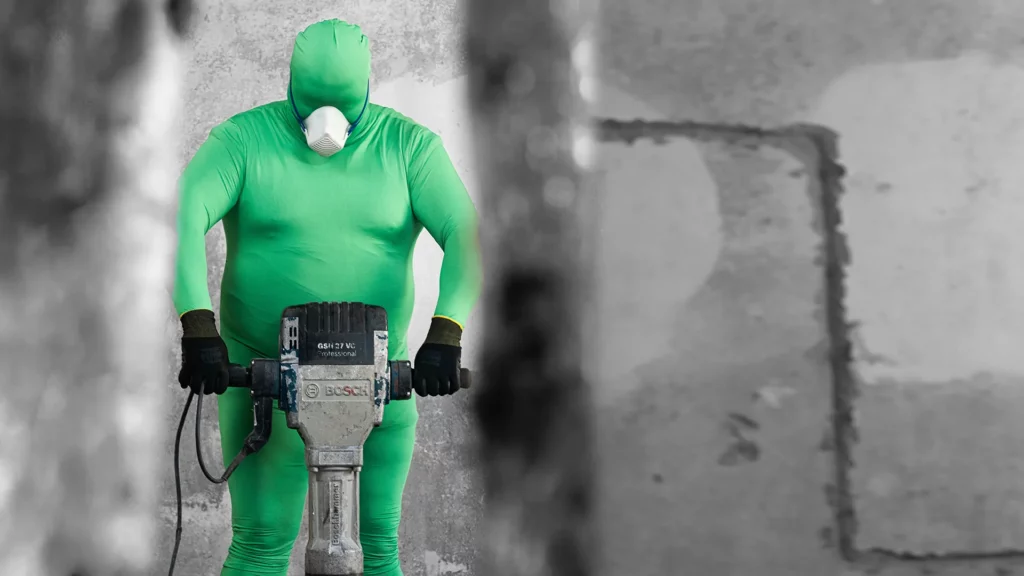 Person im grünen Ganzkörperanzug mit Staubmaske hält einen Abbruchhammer vor einer Betonwand