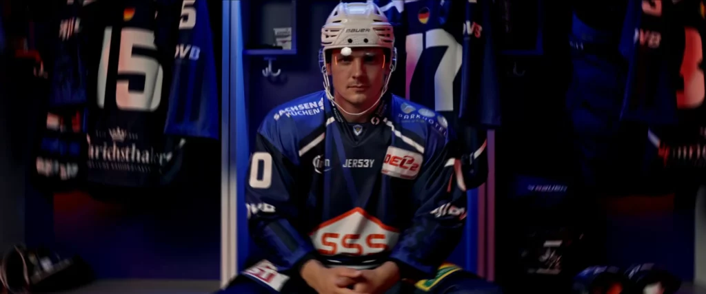 Eishockeyspieler in blauem Trikot mit Helm sitzt in der Umkleide zwischen aufgehängten Trikots