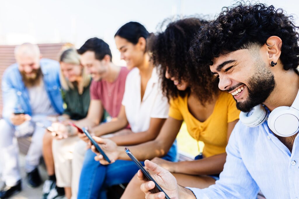 Das Bild zeigt eine Gruppe junger Menschen, die nebeneinander sitzen und gemeinsam auf ihre Smartphones schauen. Sie lachen und wirken fröhlich, was auf eine positive und entspannte Atmosphäre hinweist. Die Gruppe ist vielfältig und zeigt unterschiedliche Menschen in lässiger Freizeitkleidung, was ein Gefühl von Gemeinschaft und moderner Vernetzung vermittelt. Einige tragen Kopfhörer um den Hals, was darauf hinweist, dass sie auch Musik oder andere Medien auf ihren Geräten nutzen. Die Szene findet im Freien statt, vermutlich in einer städtischen Umgebung an einem sonnigen Tag.