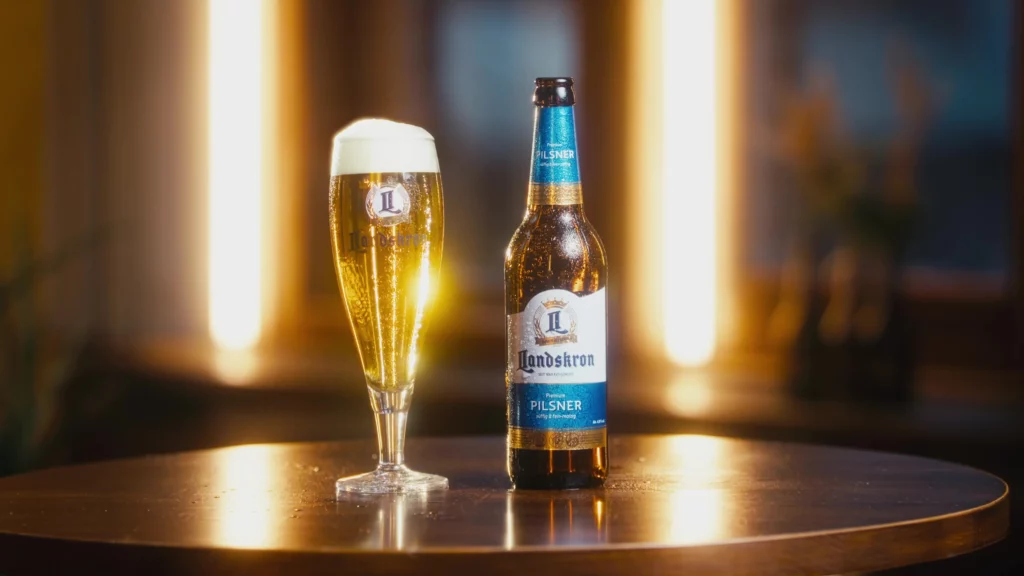 Flasche Landskron Pilsner neben einem gefüllten Bierglas auf einem Holztisch im warmen Gegenlicht