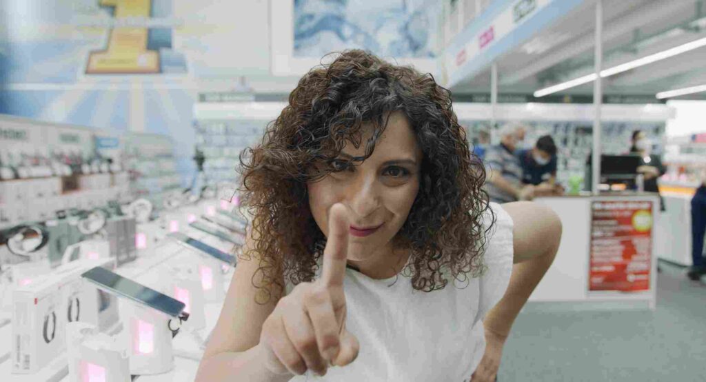 Frau zeigt lächelnd in die Kamera, dahinter die Verkaufsfläche eines Fachmarkts